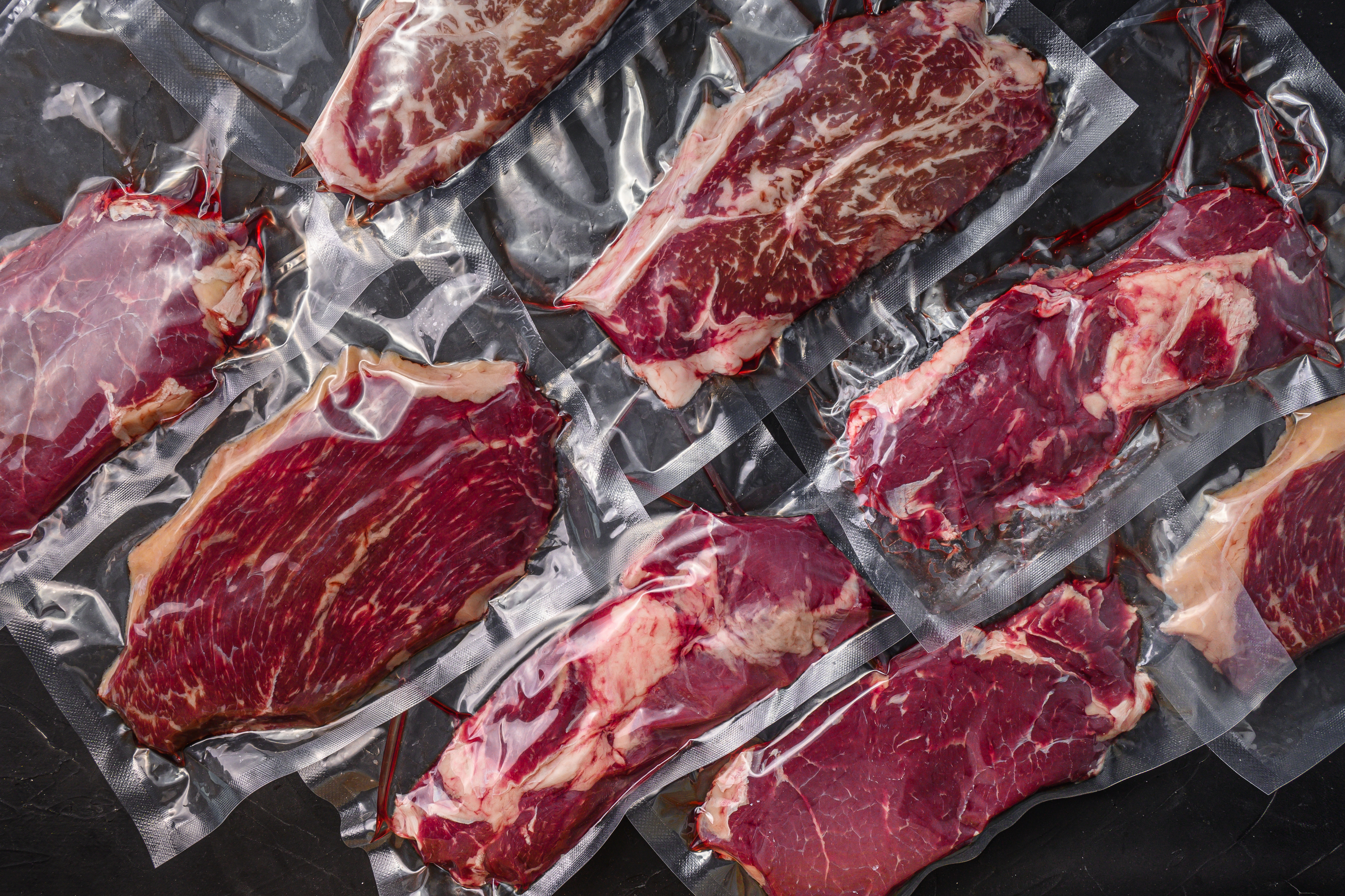 Set  of  vacuum packed organic raw beef alternative cuts: top blade, rump, picanha, chuck roll steaks, over black textured background, top view